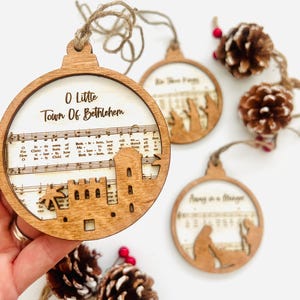 Pode incluir: Enfeites de Natal de madeira com um design rústico. Cada enfeite apresenta um design recortado com um tema religioso, incluindo partituras e os textos "O Little Town Of Bethlehem", "We Three Kings" e "Away in a Manger".