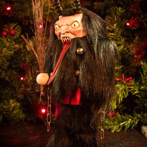 May include: A Krampus figurine with black fur, two curved horns, and a long red tongue. The figure has yellow eyes, sharp teeth, and holds a bundle of twigs. It wears a black coat with small metal bells and stands on a red and white base.