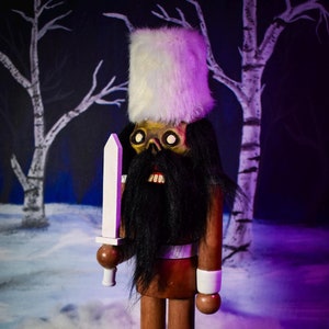 May include: A wooden nutcracker with a white fur hat and beard, holding a white sword. The nutcracker is standing on a wooden base and has a brown body with white fur boots.