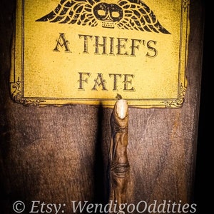 Pode incluir: Placa de madeira com o texto "A Thief's Fate" em estilo vintage. A placa apresenta um design de caveira e asas e uma pega em forma de dedo. O fundo é de madeira escura.