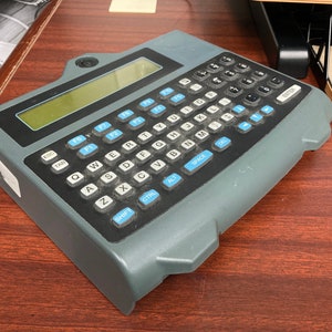 Puede incluir: Un ordenador de mano gris con un teclado azul y negro. El teclado tiene teclas etiquetadas con letras, números y teclas de función. El ordenador tiene una pequeña pantalla en la parte superior.