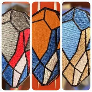 Puede incluir: Tres parches bordados con diseños geométricos. El primer parche presenta secciones grises, rojas y azules. El segundo parche tiene secciones naranjas y azules. El tercer parche muestra secciones azules y beige. Cada parche tiene un borde negro.