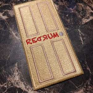 May include: A gold door-shaped patch with the word "REDRUM" embroidered in red.