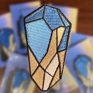 Puede incluir: Un parche bordado con forma de cristal, con colores azul y marrón. El cristal está formado por muchas formas geométricas más pequeñas.