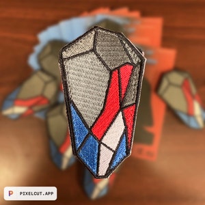Puede incluir: Un parche bordado con forma de cristal, con colores rojo, blanco y azul. El parche está hecho de tela y tiene una superficie texturizada.