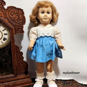 May include: A vintage doll with blonde hair and blue eyes, wearing a white eyelet top, a blue skirt, white socks, and white shoes. The doll is standing in front of a wooden clock.