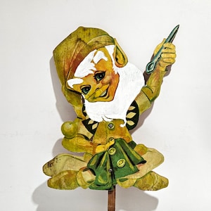 May include: A whimsical, hand-painted, wooden gnome figurine with a green hat, white beard, and a green skirt. The gnome is holding a small, silver-colored object in its right hand and is sitting on a wooden stake.