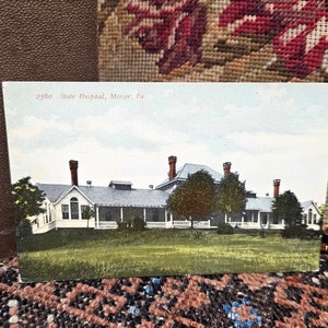 May include: Vintage postcard showing a white building with a red roof and multiple chimneys. The building is surrounded by trees and grass. The text on the postcard reads "2500 State Hospital, Mercer, Pa."