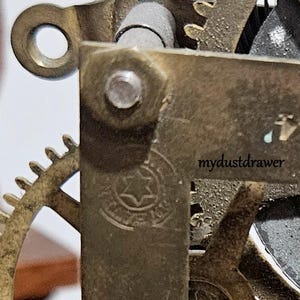 May include: Close-up of a clock mechanism with brass gears and metal components. The image shows a detailed view of the inner workings, including a gear with a star-shaped emblem and the text "mydustdrawer".