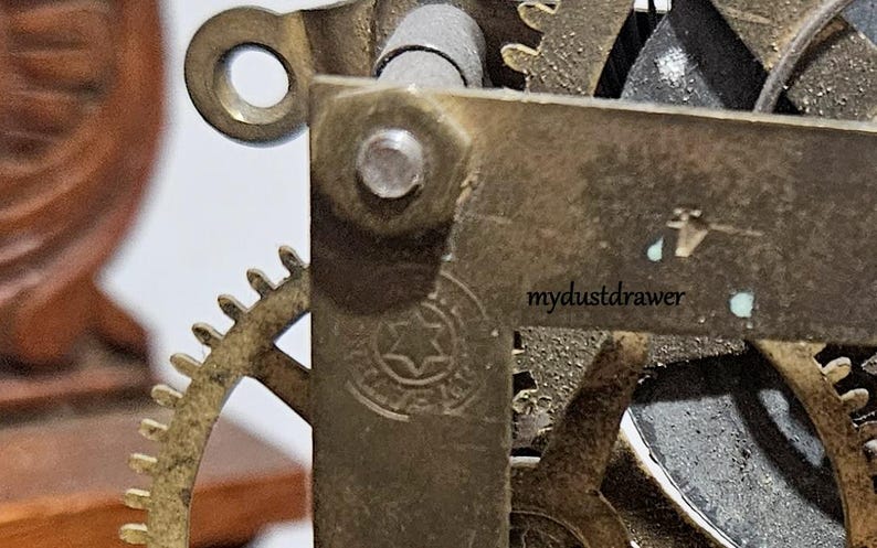 May include: Close-up of a clock mechanism with brass gears and metal components. The image shows a detailed view of the inner workings, including a gear with a star-shaped emblem and the text "mydustdrawer".