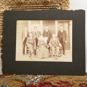 May include: A black and white vintage photograph of a family posed on a porch. Seven people are standing and sitting on chairs. The family is dressed in formal clothing. The photo is framed with a black mat.