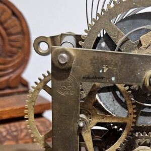 May include: Close-up of a vintage clock mechanism with brass gears and a wooden decorative element. The gears are intricately designed, showing the inner workings of the clock. The wooden element has a carved floral design.