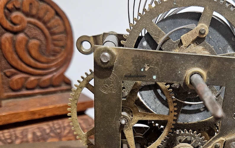May include: Close-up of a vintage clock mechanism with brass gears and a wooden decorative element. The gears are intricately designed, showing the inner workings of the clock. The wooden element has a carved floral design.