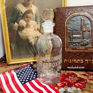 May include: A clear glass bottle with a glass stopper. The bottle is labeled "Ammonium Hydroxide NH4OH". The bottle is sitting on a table with a red, white, and blue American flag and a brown leather-bound book with Hebrew text.