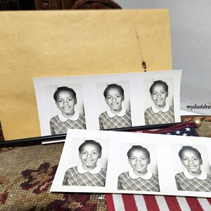 May include: Black and white photo booth strips featuring a young person with a collared shirt. The photos are arranged with a large manila envelope in the background. A small American flag is visible.