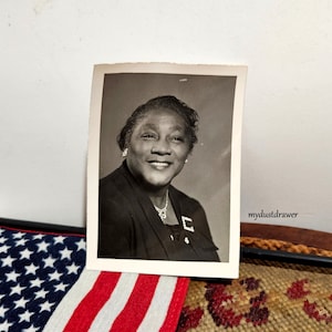 May include: Black and white photograph of a woman wearing a dark suit jacket and a pearl necklace. She is smiling and looking at the camera. The photo is printed on a white card.