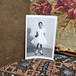 May include: Black and white photograph of a young girl wearing a white dress and a white hat. She is holding a small purse and is standing in front of a plain background.