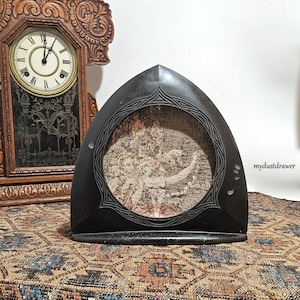 May include: A black, arched picture frame with a detailed, circular tapestry. The frame has an ornate, geometric border. An antique wooden clock is visible in the background, adding to the vintage aesthetic.