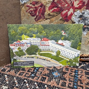 May include: Vintage postcard showing an aerial view of St. Helena Sanitarium and Hospital in Sanitarium, California. The postcard features a large white building with red roof tiles surrounded by trees.