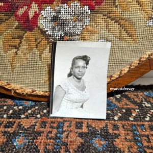 May include: Black and white photograph of a woman wearing a white patterned dress and a pearl necklace. The photo is framed and sitting on a patterned surface.