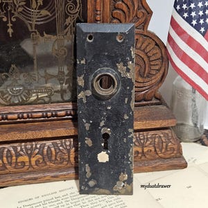 May include: A rectangular black metal door plate with a distressed finish revealing brass-colored patches. It features a central circular knob opening, a keyhole below, and four small screw holes at the corners. The surface shows significant wear.