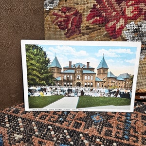 May include: A vintage postcard featuring the Main Building of the State Normal School in Lock Haven, PA. The postcard shows a large brick building with multiple turrets and a crowd of people on the lawn. The sky is blue with some clouds.