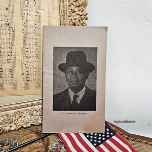 May include: Black and white photo of a man wearing a suit and a hat. The photo is on a piece of paper with the text "J. S. SIMMONS, Moderator" printed below the photo.