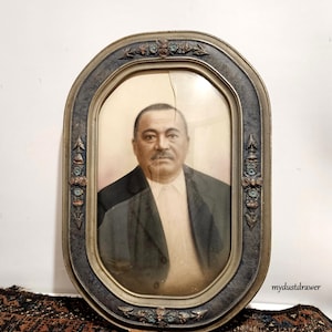 May include: A vintage oval-shaped picture frame with a gold-toned finish and ornate details. The frame holds a black and white photograph of a man in a suit.