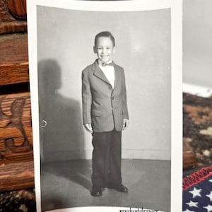 May include: Black and white photograph of a young boy wearing a suit and bow tie. He is smiling and looking at the camera.