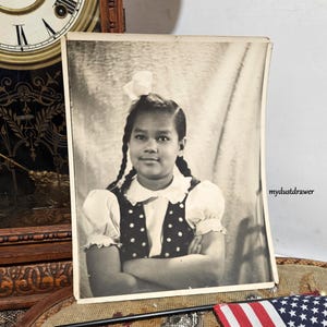 May include: Black and white vintage photograph of a young girl with braided hair, wearing a white blouse with a polka dot vest. A white bow adorns her hair. The photograph is slightly aged.