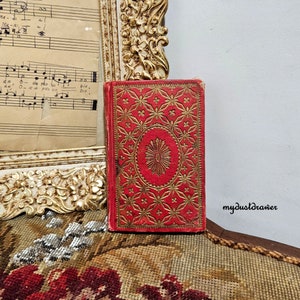 May include: A red hardcover book with gold embossed details and a floral pattern. The book is closed and sitting on a patterned surface.