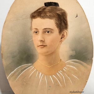 May include: An antique oval portrait of a woman with brown hair styled in a bun. She wears a gold necklace and a light-colored dress. The portrait has a faded appearance with visible wear and tear, and the text "mydustdrawer" is visible.