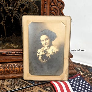 May include: A vintage photograph of a woman in a dark hat holding a bouquet of white flowers. The photo is framed in a simple cardboard frame.