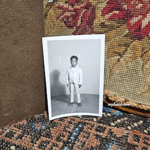 May include: Black and white photograph of a young child wearing a light-colored suit with a bow tie. The child is standing in front of a plain background. The photograph is displayed on a patterned surface.