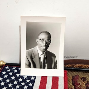 May include: Black and white photograph of a man wearing a suit and tie. He is smiling and wearing glasses. The photo is in a white frame.