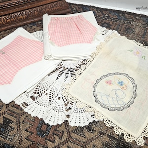 May include: A collection of vintage textiles, including white handkerchiefs with embroidered floral designs and lace trim, and pink gingham fabric. The items are arranged on a patterned rug, showcasing a nostalgic aesthetic.