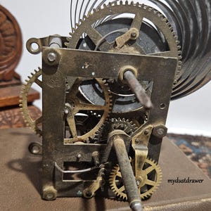 May include: Close-up of a vintage clock mechanism with intricate gears and a spiral spring. The metal components show signs of age and wear, with a mix of brass and steel elements. The image is set against a neutral background, highlighting the mechanical details.