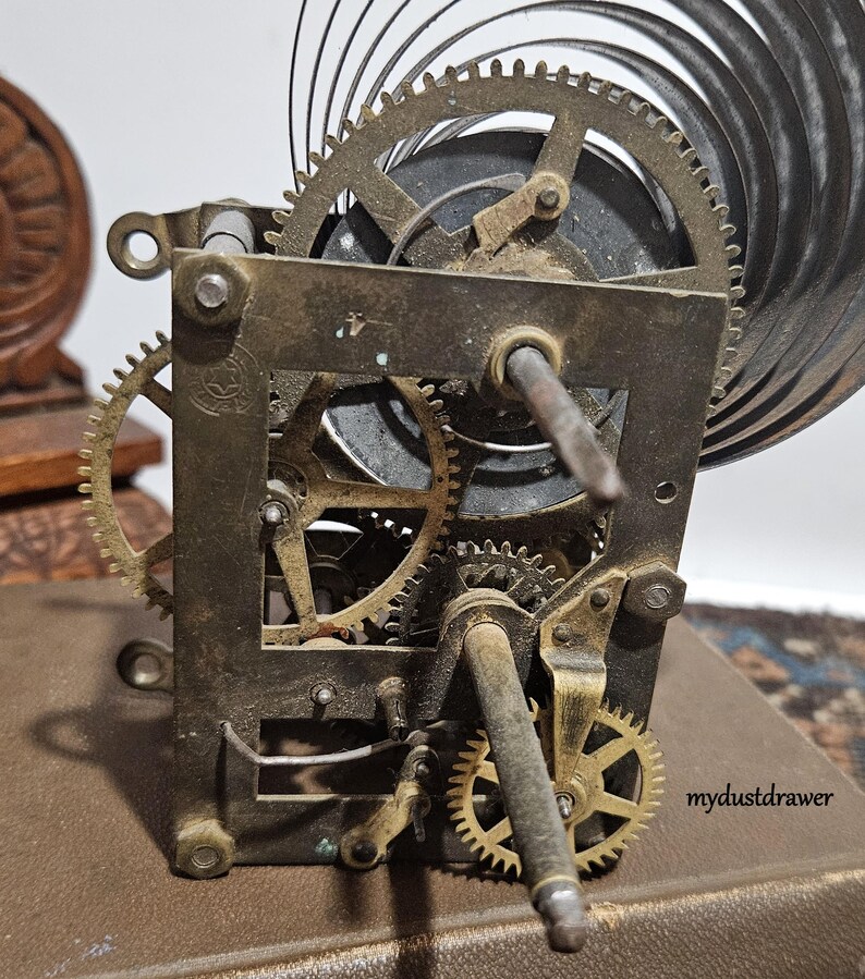 May include: Close-up of a vintage clock mechanism with intricate gears and a spiral spring. The metal components show signs of age and wear, with a mix of brass and steel elements. The image is set against a neutral background, highlighting the mechanical details.