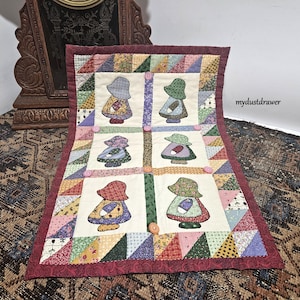 May include: A white quilted wall hanging with a red border features six colorful appliqued figures of girls wearing bonnets. The figures are arranged in a grid pattern with a button in each square. The background fabric is a patchwork of colorful fabrics.