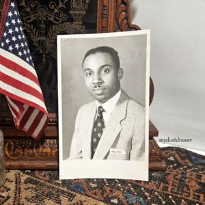 May include: Black and white photograph of a man in a suit and tie. The man is wearing a light gray suit jacket, a white shirt, and a patterned tie. An American flag is visible in the background.