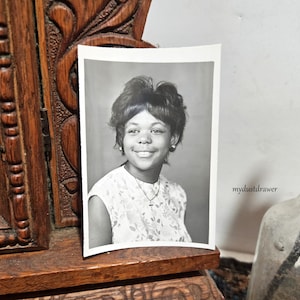 May include: Black and white photograph of a smiling person with dark hair, wearing a floral patterned top and a cross pendant necklace. The photo is displayed on a wooden surface, with a carved wooden frame visible on the left.