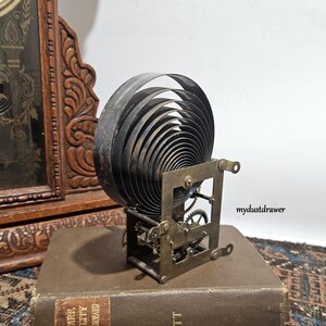 May include: A vintage clock spring mechanism with a large, coiled spring and visible gears. The metal components have a dark, aged appearance. The mechanism is set on a book, with a wooden clock frame in the background.