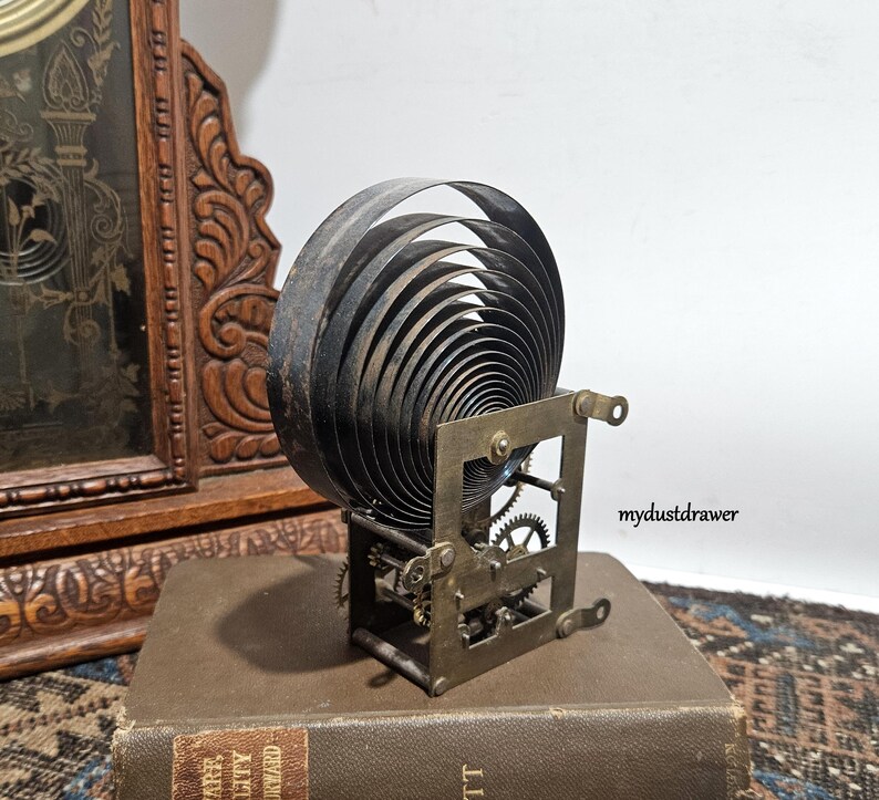 May include: A vintage clock spring mechanism with a large, coiled spring and visible gears. The metal components have a dark, aged appearance. The mechanism is set on a book, with a wooden clock frame in the background.
