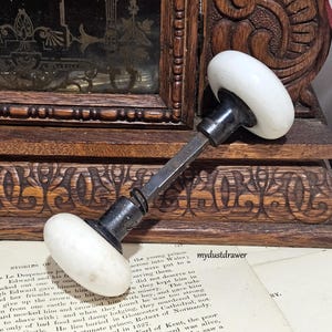 May include: A vintage door knob set featuring two smooth, round, white porcelain knobs connected by a central square metal spindle. The hardware includes dark metal collars at the base of each knob, with a small coiled spring visible on the spindle.