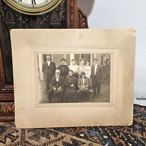 May include: A black and white vintage photograph of a family group portrait. The family is standing in front of a building with a porch. The family members are dressed in formal clothing. The photo is framed in a simple, brown frame.