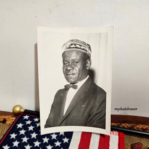 May include: Black and white photograph of a man wearing a patterned hat and a suit with a bow tie.
