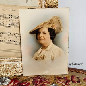 May include: A vintage postcard featuring a black and white photo of a woman wearing a large, wide-brimmed straw hat with a pink flower accent. The woman is looking directly at the camera and has a gentle smile. The background is a light blue.