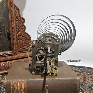 May include: A vintage clock movement with visible gears and a coiled spring. The mechanism is set on a brown book with gold lettering. A carved wooden clock frame is visible in the background.