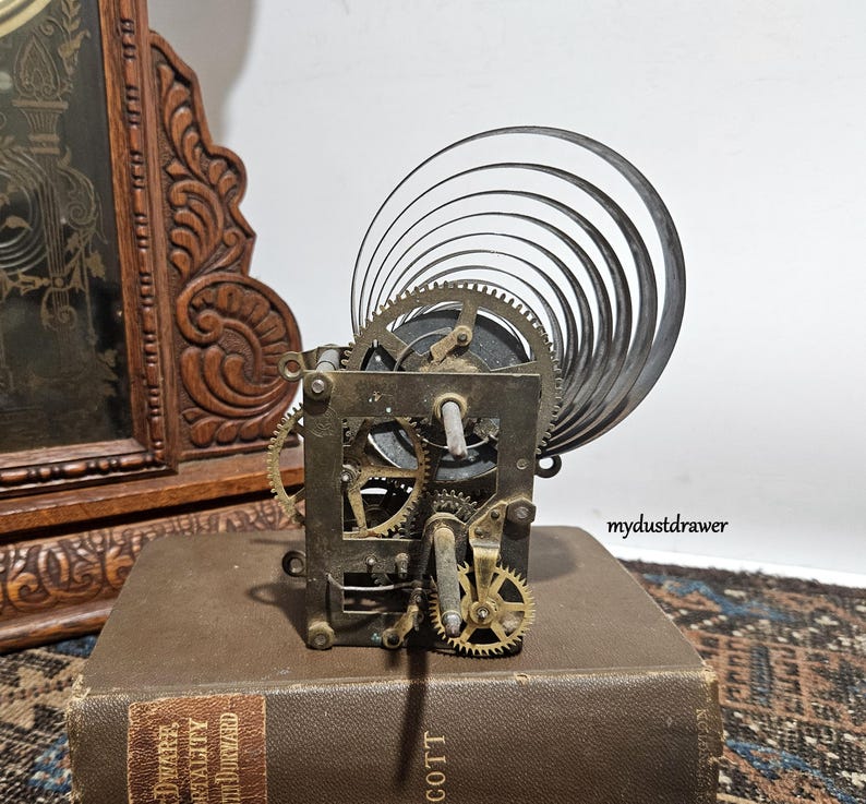 May include: A vintage clock movement with visible gears and a coiled spring. The mechanism is set on a brown book with gold lettering. A carved wooden clock frame is visible in the background.