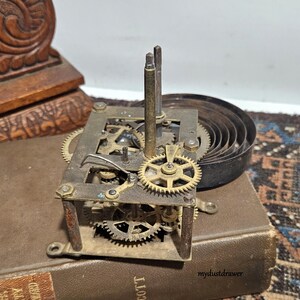 May include: Close-up of a vintage clock movement with brass gears, springs, and metal frame. The clock mechanism is set on a brown book. The image shows the intricate details of the clock's internal components.